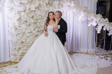 Bir gelin ve damat beyaz bir duvarın önünde poz veriyorlar. Gelin beyaz bir elbise giyiyor ve damat siyah bir takım elbise giyiyor. Sahne mutlu ve kutlama dolu.