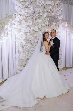 Bir gelin ve damat beyaz bir duvarın önünde poz veriyorlar. Gelin beyaz bir elbise giyiyor ve damat siyah bir takım elbise giyiyor. Sahne mutlu ve kutlama dolu.