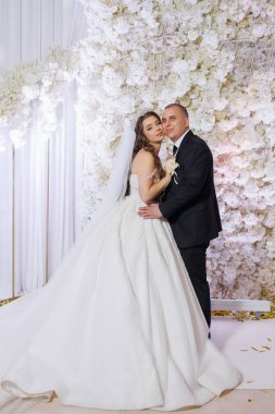 Bir gelin ve damat beyaz bir duvarın önünde poz veriyorlar. Damat siyah bir takım giyerken gelin beyaz bir elbise ve duvak takıyor. Sahne mutlu ve kutlama dolu.