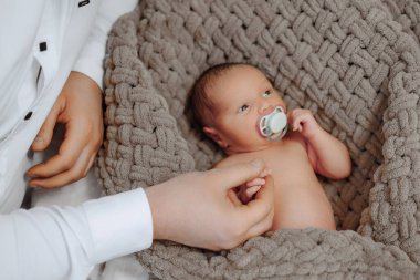 Ağzında emzik olan bir bebek battaniyenin üzerinde yatıyor. Bebek bir insan tarafından tutuluyor.