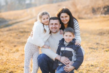 Tarlada fotoğraf çektirmek için poz veren dört kişilik bir aile. Baba beyaz bir kazak giyiyor ve çocuklar ceket giyiyor. Sahne sıcak ve mutlu.