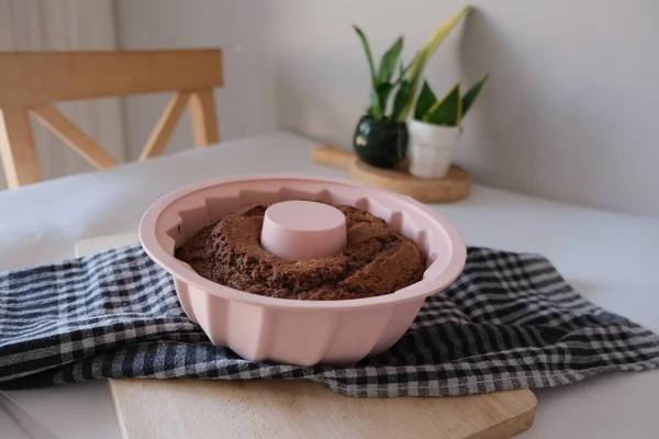 Baked cake in a silicone mold, baked cake with cacao in a pink round silicone mold on dark colored checked kitchen cloth on a white kitchen table. The concept photo of homemade food.