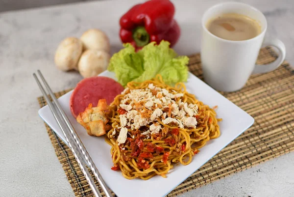 Mie Goreng, baharatlı kızarmış şehriye, tavuklu ve biberli. Yanında da bir fincan kahve ve sebze ve gri arka planda servis ediliyor.