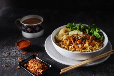 Mie ayam, tavuklu erişte ve sebzeler beyaz kasede, Endonezya geleneksel yemekleri koyu ve dokuma arka planda. Kızarmış soğan ve bir fincan kahveyle servis edilir.