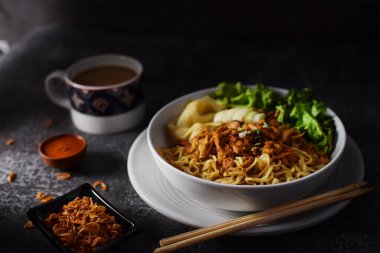 Mie ayam, tavuklu erişte ve sebzeler beyaz kasede, Endonezya geleneksel yemekleri koyu ve dokuma arka planda. Kızarmış soğan ve bir fincan kahveyle servis edilir.