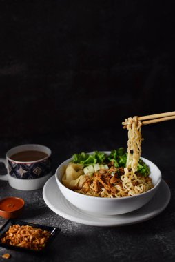 Mie ayam, tavuklu erişte ve sebzeler beyaz kasede, Endonezya geleneksel yemekleri koyu ve dokuma arka planda. Kızarmış soğan ve bir fincan kahveyle servis edilir.
