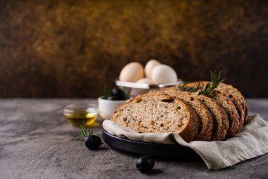 Bir ızgarada kepekli tahıllı un ve kabak tohumlarından taze pişmiş ekşi ekmek, kırsal bir ahşap masada zeytinyağı ve siyah zeytin. Esnaf ekmeği.