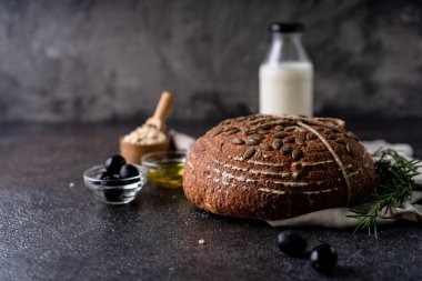 Bir ızgarada tam tahıllı un ve balkabağı tohumlarından fırınlanmış ekşi ekmek, kırsal bir ahşap masada zeytinyağı ve siyah zeytin. Esnaf ekmeği.