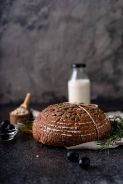 Bir ızgarada tam tahıllı un ve balkabağı tohumlarından fırınlanmış ekşi ekmek, kırsal bir ahşap masada zeytinyağı ve siyah zeytin. Esnaf ekmeği.