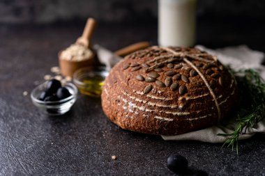 Bir ızgarada tam tahıllı un ve balkabağı tohumlarından fırınlanmış ekşi ekmek, kırsal bir ahşap masada zeytinyağı ve siyah zeytin. Esnaf ekmeği.