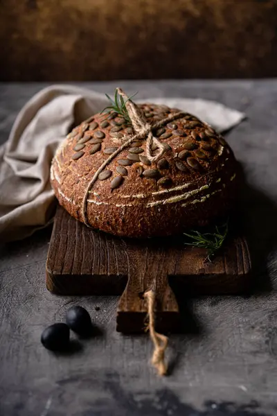 Bir ızgarada tam tahıllı un ve balkabağı tohumlarından fırınlanmış ekşi ekmek, kırsal bir ahşap masada zeytinyağı ve siyah zeytin. Esnaf ekmeği.