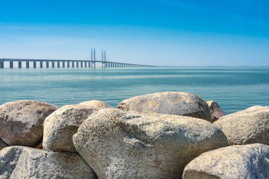Öresund Köprüsü yazın. Mavi gökyüzü ve mavi su. Ön planda taşlar var. Köprü İsveç ile Danimarka arasında. Öresund bağlantısının köprüleri Saltholm 'un güneyindeki Pepparholm Adası' ndan Limhamn 'ın güneyindeki Lernacken Adası' na kadar uzanıyor..