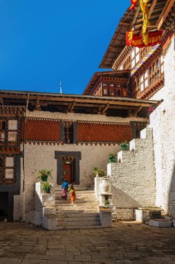 Tow girls in traditional clothes walking up the stairs in Trongsa dzong inner courtyard. clipart