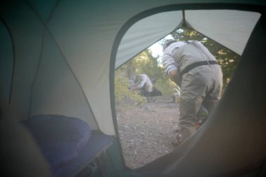 Kamp alanındaki çadırın içindeki fermuarlı kapaktan bakın. İki adam günlük aktivitelere hazırlanıyor.