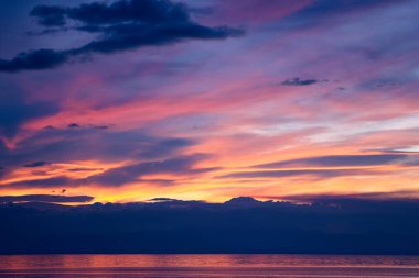 Görkemli turuncu ve mor gün batımı siluetli dağların üzerinde ve sakin su parlak ışığı yansıtıyor.