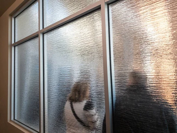 Couple queuing behind textured a frosted glass window or partition standing with their back to the camera at an oblique angle