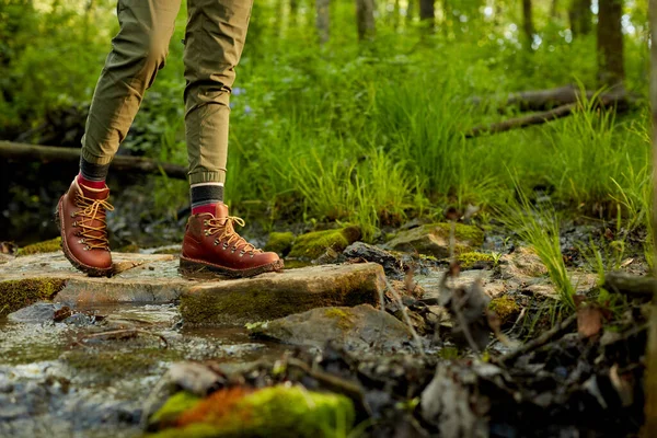 stock image Woman hiking across a small rocky stream bordered by lush green grass in a close up view of her brown leather boots