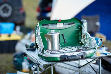 Paslanmaz çelik kahve demliği, küçük taşınabilir bir kamp gaz ocağında ısıtılıyor. Dışarıdaki katlama masasında kahvaltı hazırlıyor.