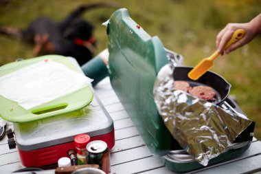 Adam, elinde et ve spatulayı yakından gören, küçük taşınabilir bir portatif üzerinde, kamp gaz ocağında çiğ hamburger köftesi pişiriyor.