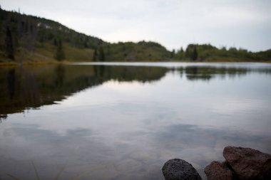 Kasvetli bulutlu bir günde, sessiz sakin bir göl. Gri bulutlar ve etrafındaki tepeler suya yansıyor.