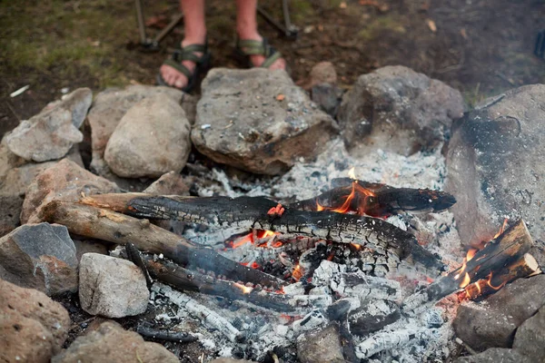 Camper standing alongside a burning campfire with glowing hot coals from logs of wood surrounded by small rocks as he waits to begin cooking a meal