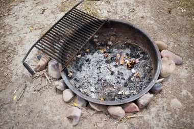 Bir kamp ateşini yakmak için boş metal tepsi. Yerde taşlarla dengelenmiş kül kalıntıları ve yemek pişirmek için bir ızgara.