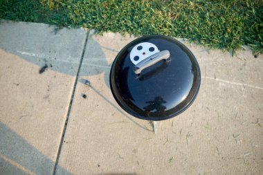 Yeşil bir çimenliğin yanında asfalt bir geçitte kapağı kapalı duran küçük küresel taşınabilir bir barbekü manzarası