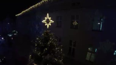 Karlı bir kasaba meydanının ortasında güzel bir Noel ağacı duruyor. Parlayan beyaz ışıklarla süslenir ve üzerinde büyük, parıldayan bir yıldız bulunur. Ağaç karla kaplı ağaçlarla çevrili.