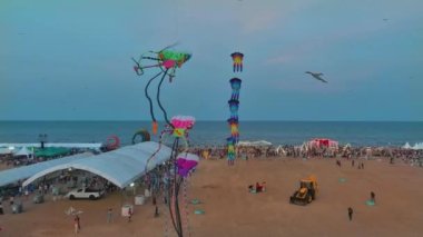 Bir plaj festivalinin büyüleyici hava manzarası, gökyüzünde çeşitli şekil ve boyutlarda renkli uçurtmalar süzülüyor. Kumsal şenliklerin tadını çıkaran insanlarla dolu.