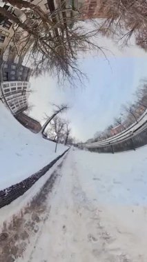 Bu bir şehir parkında karlı bir yolda 360 derecelik bir video turu. Yol karla kaplı ve her iki taraftaki ağaçlar ve çalılar çıplak. Yolun sağında bir çit var. Gökyüzü...