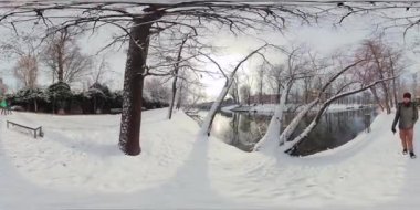 Bu 360 derecelik videoda karlı bir parkta bir yürüyüş yap. Video, karla kaplı bir parkın görüntüsüyle başlıyor. Kar el değmemiş ve el değmemiş, ağaçlar da çıplak. Bir adam yürüyor.