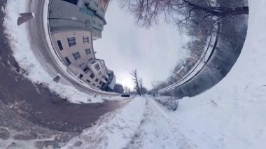 Bu, şehir parkındaki karlı bir yolun 360 derecelik bir videosu. Yol ağaçlar ve çalılarla çevrili ve sağda bir çit var. Uzakta bir bina var. Zemin kaplanmış.