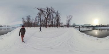 Bu 360 derecelik video, karlı bir parkta yürüyen bir adamı gösteriyor. Adam siyah bir ceket ve kot pantolon giyiyor ve ılımlı bir tempoda yürüyor. Park karla kaplı ve başka bir şey yok.