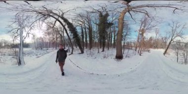 Bu, karlı bir kış gününde karlı bir ormanda yürüyen bir adamın 360 derecelik görüntüsü. Adam siyah bir ceket ve kot pantolon giyiyor ve kahverengi bir çanta taşıyor. Karla kaplı bir yolda yürüyor.