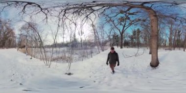 Bu, karlı bir ormanda yürüyen bir adamın 360 derecelik görüntüsü. Adam siyah bir ceket, kot pantolon ve siyah bir sırt çantası giyiyor. Karla kaplı bir yolda yürüyor, uzun ağaçlarla çevrili.