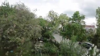 Video, arka bahçesi, çimleri, çalıları ve ağaçları olan bir arka bahçeyle açılıyor. Kamera sola dönüyor, bej tuğlalı ve geniş pencereli bir apartmanı gösteriyor. Kamera devam ediyor.