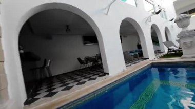 Video beyaz duvarlar ve yemyeşil yeşilliklerle çevrili havuzlu güzel bir avlu gösteriyor. Avluda mozaik kiremit tabanlı büyük, mavi bir yüzme havuzu bulunuyor. Havuz çevrili.
