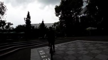 Genç bir adam, bir yaz öğleden sonra sakin bir Avrupa şehir parkında bisikletini sürüyor. Park yemyeşil çimlerle ve yemyeşil ağaçlarla dolu. Yürüyen ve banklarda oturan birkaç kişi var. Adam.