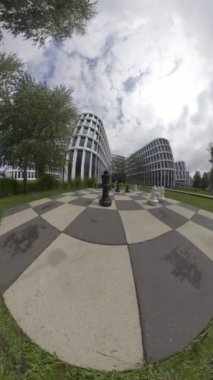 Arka planda modern ofis binaları olan çimenli bir alanda açık hava satranç tahtasının 360 derecelik bir videosu. Video satranç tahtasının etrafında döner ve parçaları farklı açılardan gösterir..