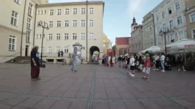 Polonya 'nın Opole kentindeki eski kasaba meydanında bir sokak sanatçısı hokkabazlık yapıyor. Oyuncu mavi bir gömlek ve kot pantolon giyiyor ve sakalı var. Üç top ile hokkabazlık yapıyor ve çok yetenekli. Kalabalık var.