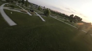 Bu FPV drone görüntüleri büyük bir çimleri, ağaçları ve taş yolu olan bir parkı gösteriyor. İnsansız hava aracı çimlerin üzerinde uçarak parkın kuş bakışı görüntüsünü sağlıyor. Görüntüler akşam çekiliyor ve güneş...