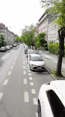 Video, bir şehrin panoramik görüntüsüyle başlıyor. Kamera soldan sağa hareket ediyor, çeşitli binaları, sokakları ve ağaçları gösteriyor. Gökyüzü, kabarık beyaz bulutlarla mavidir