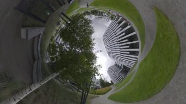 Bu balık gözü merceğiyle çekilen 360 derecelik bir video küçük bir gezegen etkisi yaratıyor. Videoda ortada ağaç olan bir park ve arka planda birkaç bina var. Gökyüzü bulutlu.