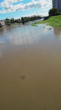 Video, bulanık suyu yavaşça akan bir nehri gösteriyor. Nehir geniştir ve kıyılarda ağaçlar ve binalar vardır. Su kirli kahverengi bir renktir ve enkazda yüzen bir sürü enkaz vardır.