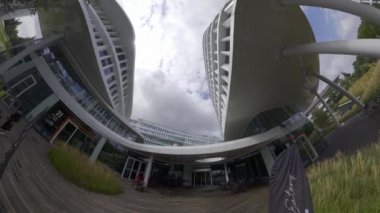 Bu, cam cepheli ve kıvrımlı girişi olan modern bir ofis binasının videosu. Bina bir şehirde yer almaktadır ve diğer yüksek binalarla çevrilidir. Video yerden alınıyor ve...