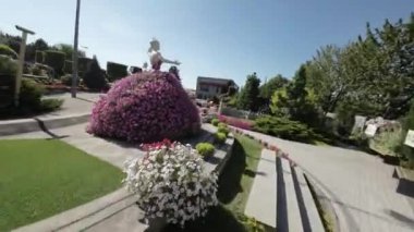 Bu, heykelleri olan güzel bir çiçek bahçesinin videosu. Bahçe pembenin ve morun çeşitli tonlarında renkli çiçeklerle dolu. Bahçede birkaç kadın heykeli var, her biri