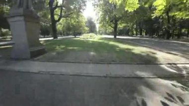 Polonya 'nın Varşova kentindeki bir parktan geçen birinci şahıs görüntüsü. Video, ağaçlarla kaplı olan park girişinin görüntüsüyle başlıyor. Kamera bir heykelin yanından geçerken bir patikadan aşağı uçar.