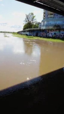 Video, şehirde bir köprünün altından akan bulanık bir nehri gösteriyor. Su kahverengi ve donuk, ve yüzeyde yüzen bir sürü enkaz var. Nehir beton duvarlarla çevrili.