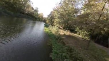 FPV 'nin nehir boyunca ve park boyunca uçuşu birinci şahıs görüntüsü. Video, insansız hava aracının perspektifinden çekildi ve manzaranın benzersiz bir görüntüsünü sağlıyor. İnsansız uçak nehir boyunca uçuyor.