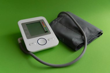 Modern dijital tonometreye yakın plan, kan basıncı kelepçeli, parlak yeşil bir arka plan üzerine kurulmuş. Beyaz cihazın gri bir ekranı var. Göstergeler ve kontrol düğmeleri var. Mavi yüzüklü olan da dahil.. 